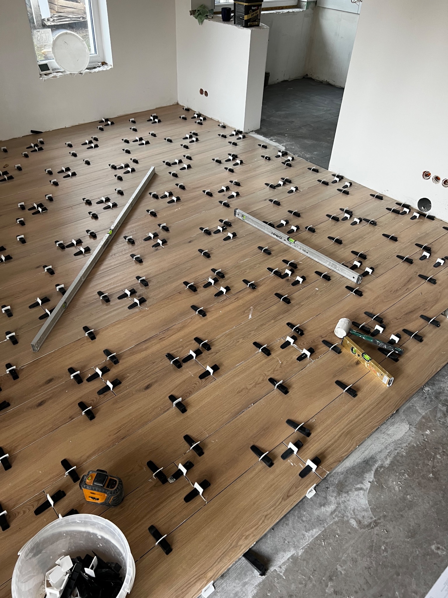 Układanie paneli podłogowych z użyciem poziomicy i klipsów dystansowych w jasnym pomieszczeniu. Widoczny fragment surowej podłogi i narzędzia pracy.