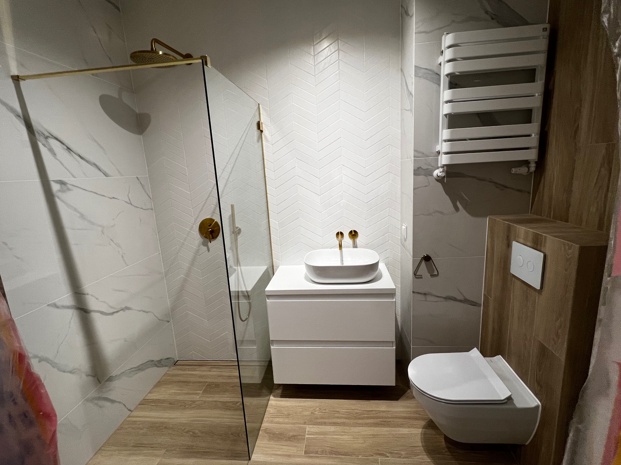 Nowoczesna łazienka z kabiną prysznicową typu walk-in, białą umywalką nablatową na szafce i grzejnikiem drabinkowym. Płytki imitujące marmur i drewno.