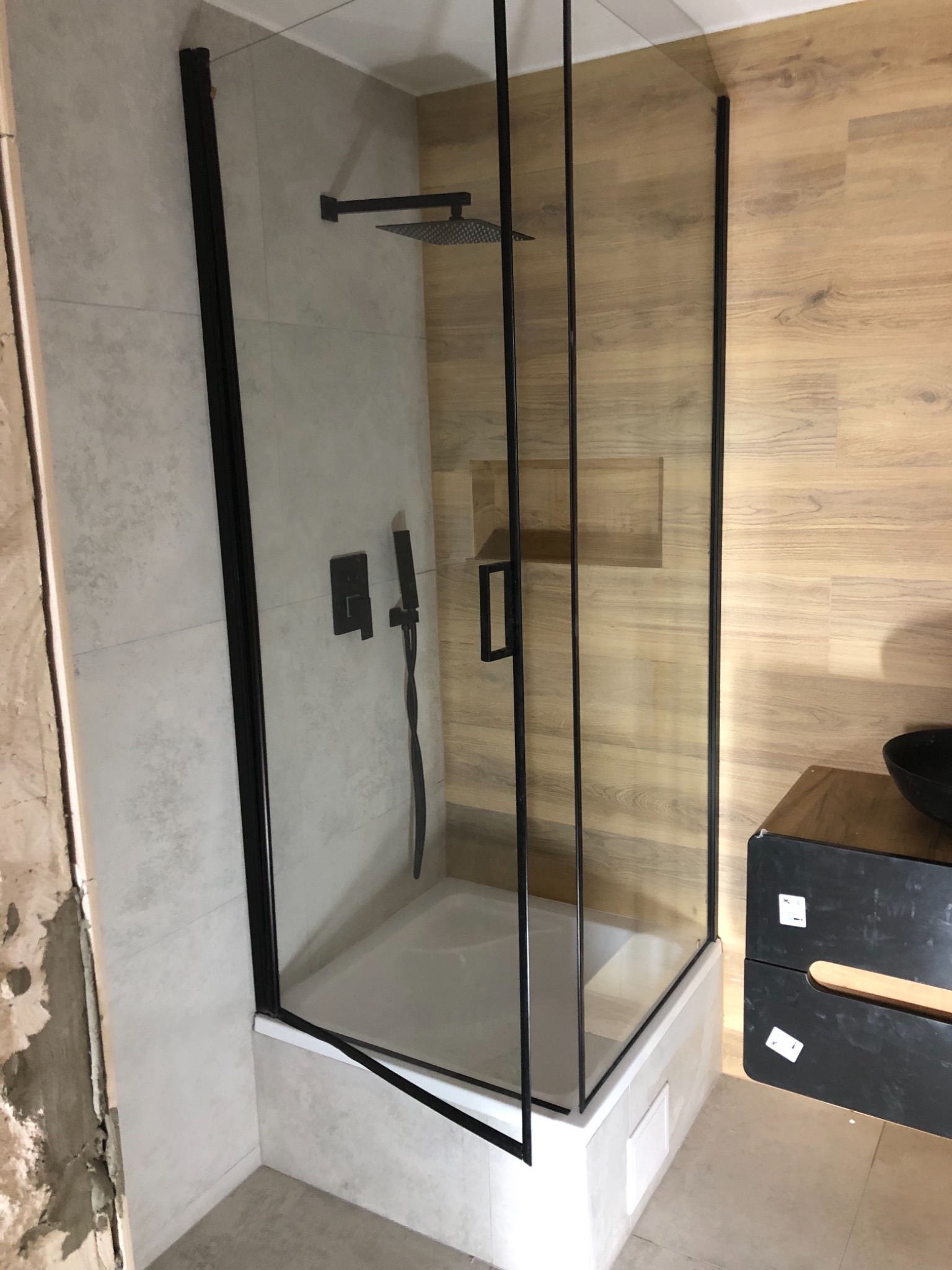 Nowoczesna kabina prysznicowa z czarnymi profilami i deszczownicą, zamontowana w łazience z wykończeniem imitującym drewno i szare płytki, widoczne ślady prac remontowych na ścianie.