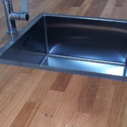 Blat z jasnego drewna z wmontowanym, prostokątnym, metalowym zlewem i baterią.