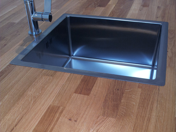 Blat z jasnego drewna z wmontowanym, prostokątnym, metalowym zlewem i baterią.