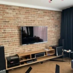 Telewizor zawieszony na ścianie z czerwonej cegły, pod nim drewniana szafka RTV z elektroniką, parkiet w jodełkę, czarne zasłony w oknie.