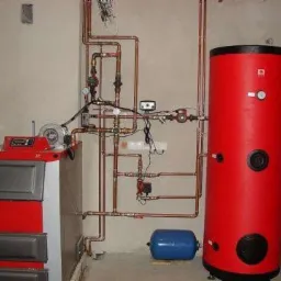 Kocioł na paliwo stałe z podajnikiem, połączony miedzianą instalacją z czerwonym zasobnikiem ciepłej wody użytkowej i niebieskim naczyniem przeponowym w pomieszczeniu kotłowni.