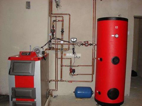 Kocioł na paliwo stałe z podajnikiem, połączony miedzianą instalacją z czerwonym zasobnikiem ciepłej wody użytkowej i niebieskim naczyniem przeponowym w pomieszczeniu kotłowni.