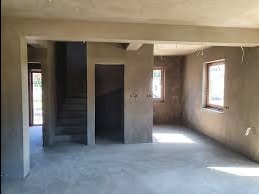 Surowe wnętrze po tynkowaniu maszynowym: ściany, sufit i podłoga pokryte świeżym, szarym tynkiem. Widoczne okna i schody. Minimalistyczny, industrialny charakter.