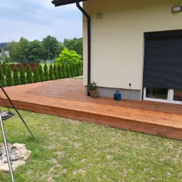 Nowoczesny taras z drewna z paleniskiem i grillem trójnożnym na trawniku, widok na dom z roletami zewnętrznymi i zielone otoczenie.