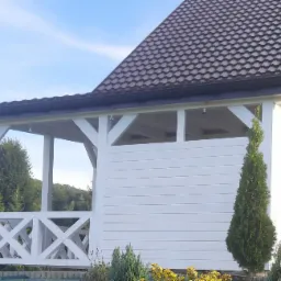 Biała altana przylegająca do domu z brązowym dachem, widok z zewnątrz, z elementami dekoracyjnymi i zielenią w otoczeniu.