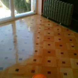 Parkiet po renowacji z wyraźnym połyskiem, widoczne odbicie światła z okna, wzór w kwadraty z ciemnymi akcentami, przy ścianie grzejnik.