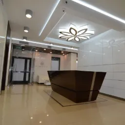 Nowoczesne wnętrze z błyszczącymi białymi szafkami w zabudowie, ciemnobrązową recepcją z metalowymi paskami i dekoracyjnym oświetleniem sufitowym w kształcie kwiatu.