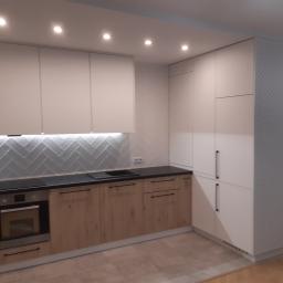 Pracownia Stolarska Skowron - Nowoczesna kuchnia z białymi górnymi szafkami i drewnopodobnymi dolnymi, czarny blat, białe płytki w jodełkę na ścianie, wysoka zabudowa z perforowanymi drzwiami po prawej stronie.