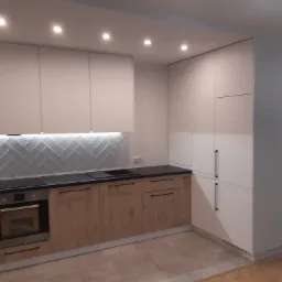 Nowoczesna kuchnia z białymi górnymi szafkami i drewnopodobnymi dolnymi, czarny blat, białe płytki w jodełkę na ścianie, wysoka zabudowa z perforowanymi drzwiami po prawej stronie.