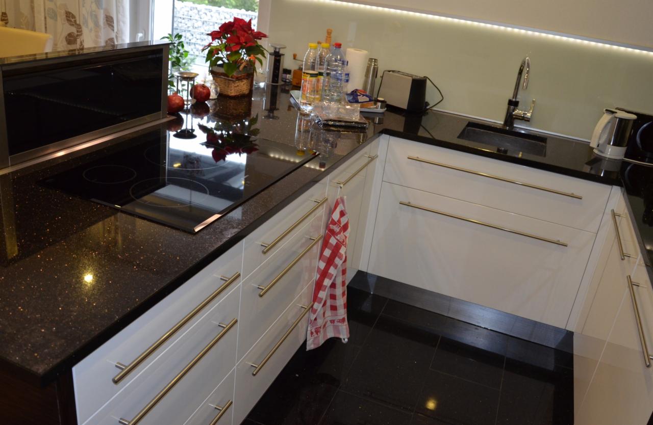 Nowoczesna kuchnia z czarnym, błyszczącym blatem z drobinkami, białymi szafkami z długimi, poziomymi uchwytami i płytą indukcyjną z wbudowanym okapem; na blacie doniczka z poinsecją i akcesoria...