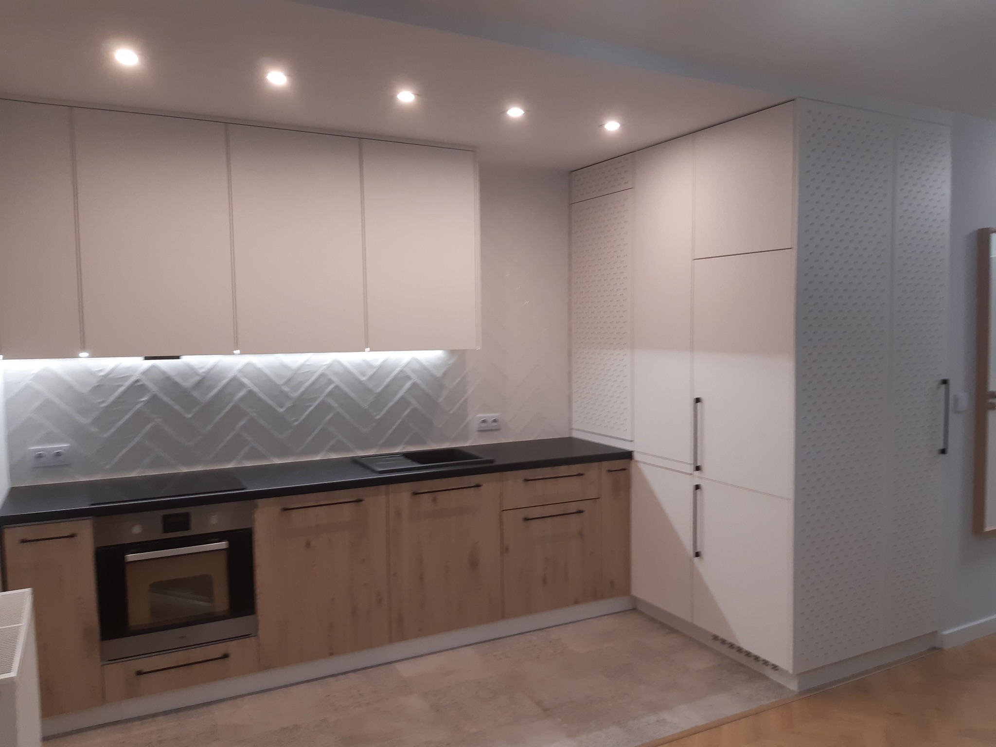 Nowoczesna kuchnia z białymi górnymi szafkami i drewnopodobnymi dolnymi, czarny blat, białe płytki w jodełkę na ścianie, wysoka zabudowa z perforowanymi drzwiami po prawej stronie.