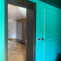 Turkusowa ściana z dekoracyjnymi panelami i czarną stolarką drzwiową, widok na drewnianą podłogę ułożoną w jodełkę i schody z czarnymi balustradami w głębi.