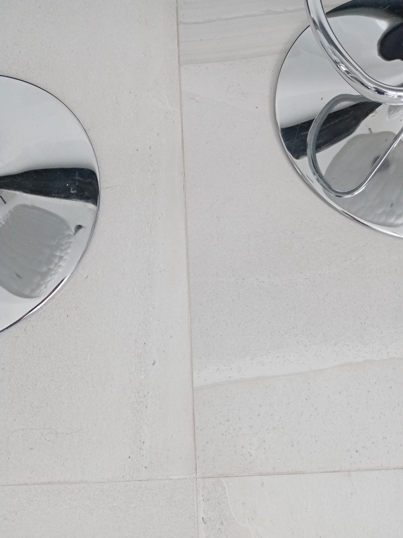 Fragment jasnej, kwadratowej podłogi z płytek, z widocznymi podstawami dwóch chromowanych krzeseł barowych.