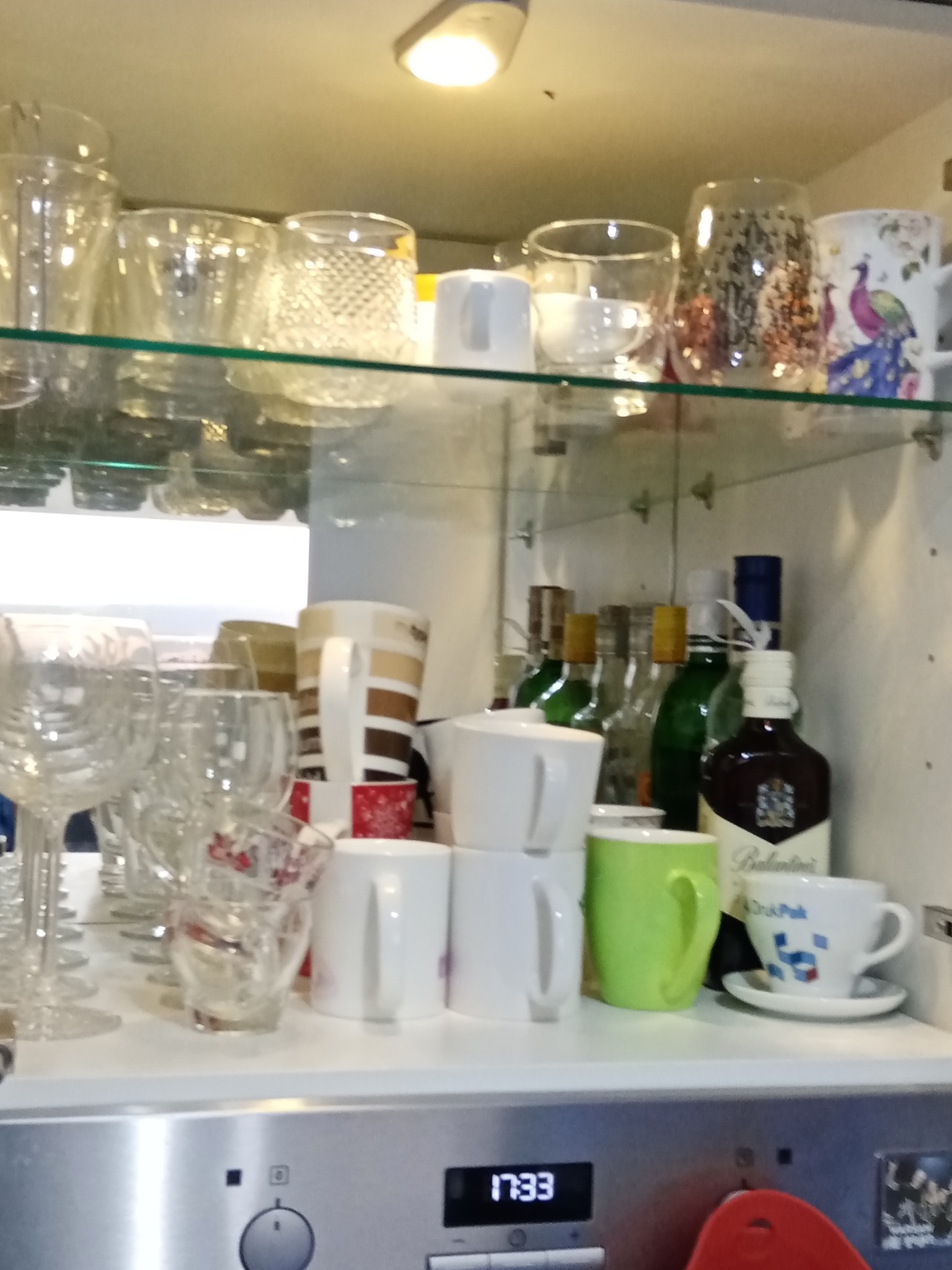 Uporządkowana szafka kuchenna z różnorodnym szkłem, filiżankami i butelkami alkoholu, widoczny wyświetlacz piekarnika z godziną 17:33.
