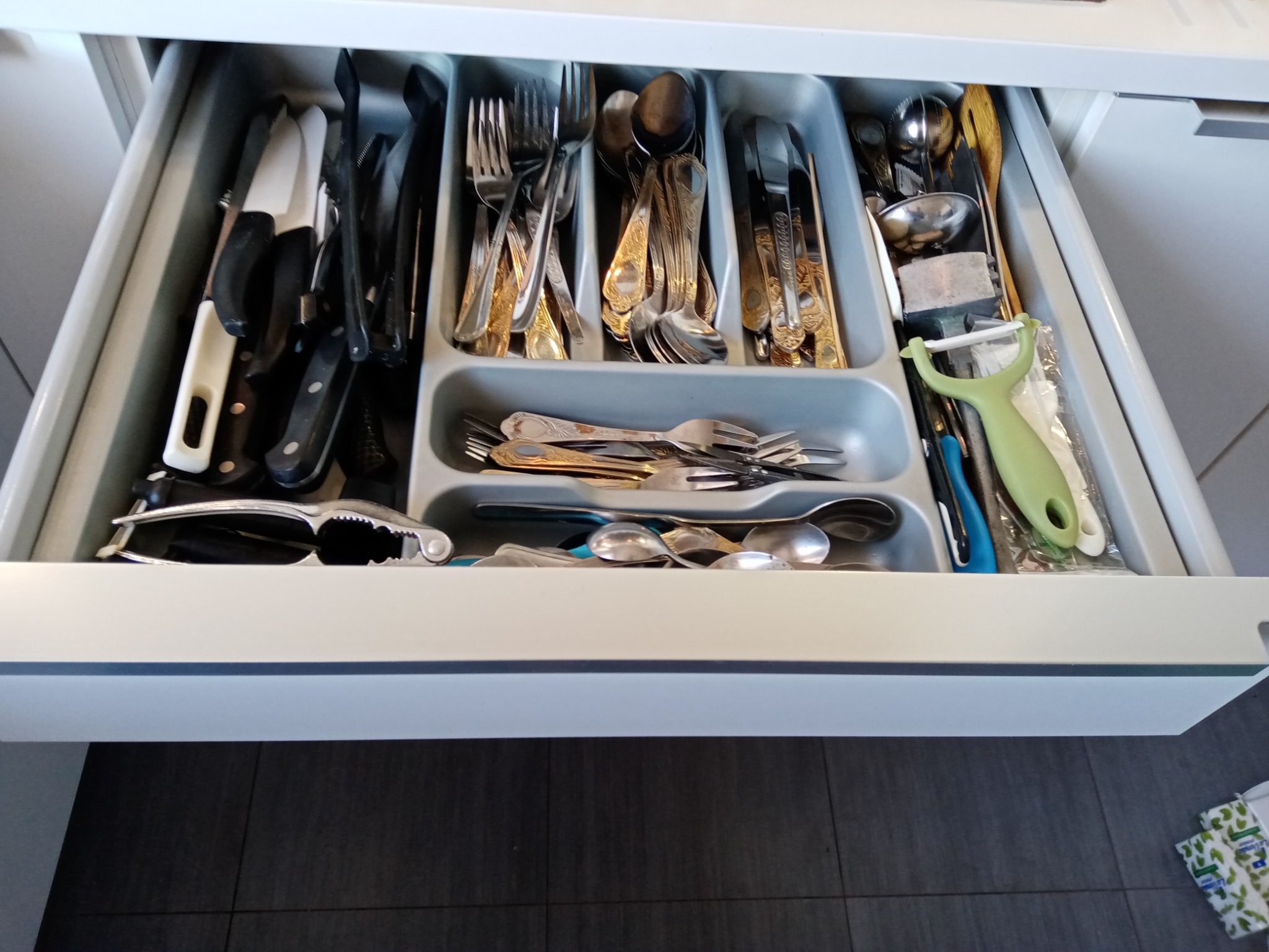 Otwarta szuflada kuchenna z organizerem na sztućce i akcesoria, widoczne noże, widelce, łyżki, obieraczka do warzyw i dziadek do orzechów.