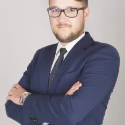 Portret mężczyzny w okularach i niebieskim garniturze, z zegarkiem i bransoletką na nadgarstku, pozującego z założonymi rękami na neutralnym tle.