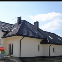 Nowoczesny dom jednorodzinny z ciemną dachówką i orynnowaniem, widoczne okna dachowe i komin z cegły, na pierwszym planie plac zabaw dla dzieci.