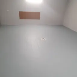 Jasnoszara, gładka podłoga w garażu widziana pod kątem, z widoczną bramą garażową po lewej stronie i kratką ściekową w posadzce.