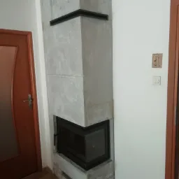 Narożny kominek z szarą obudową, czarnym wkładem i elementami dekoracyjnymi, umieszczony w rogu pomieszczenia z drewnianą podłogą i drzwiami.