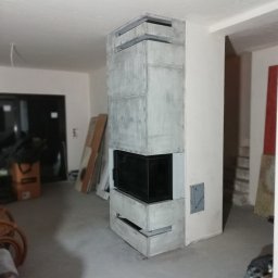 EMBUD Kominki - Nowoczesny, surowy kominek z szarego betonu architektonicznego, z czarnym wkładem, w surowym wnętrzu. Widoczne elementy instalacji i wykończenia.