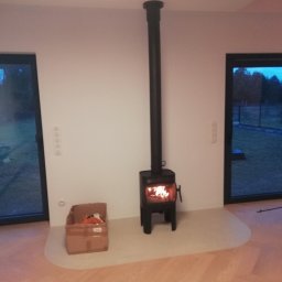 EMBUD Kominki - Czarny piec wolnostojący z rurą dymową na tle białej ściany, z płomieniami widocznymi wewnątrz. Podłoga z jasnego drewna ułożona w jodełkę.