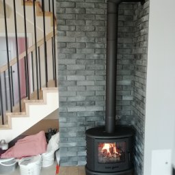 EMBUD Kominki - Czarny piec wolnostojący z palącym się ogniem, umieszczony w rogu pomieszczenia wykończonego szarą cegłą. Widoczne schody i akcesoria malarskie.