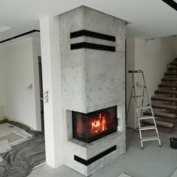 EMBUD Kominki - Narożny, obudowany kominek z paleniskiem w trakcie wykańczania. Widoczne płytki, drabina i surowe ściany. Nowoczesny design z czarnymi akcentami.