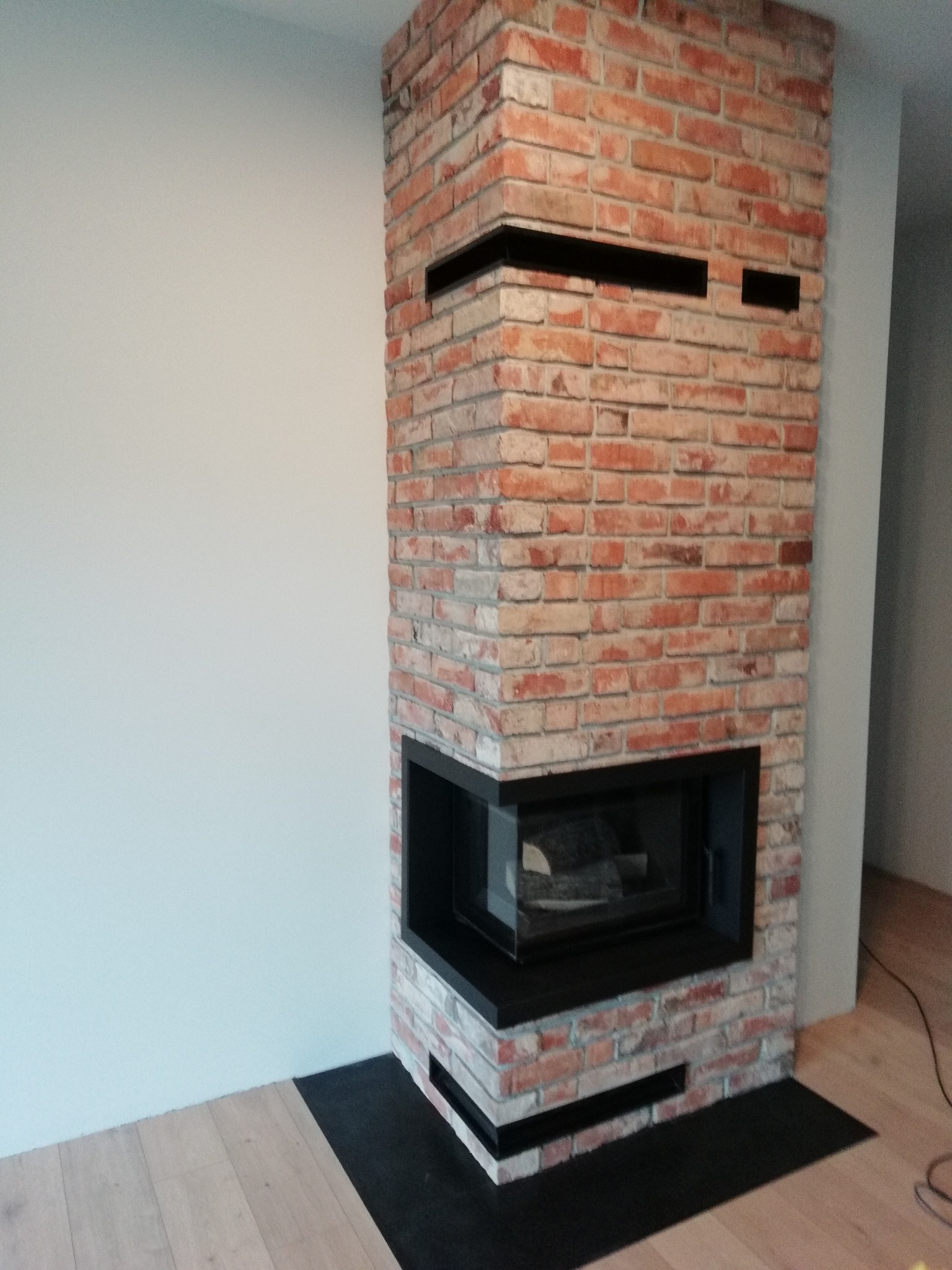 Nowoczesny wkład kominkowy z obudową z cegły klinkierowej, wykończony czarnymi elementami, na jasnej podłodze drewnianej