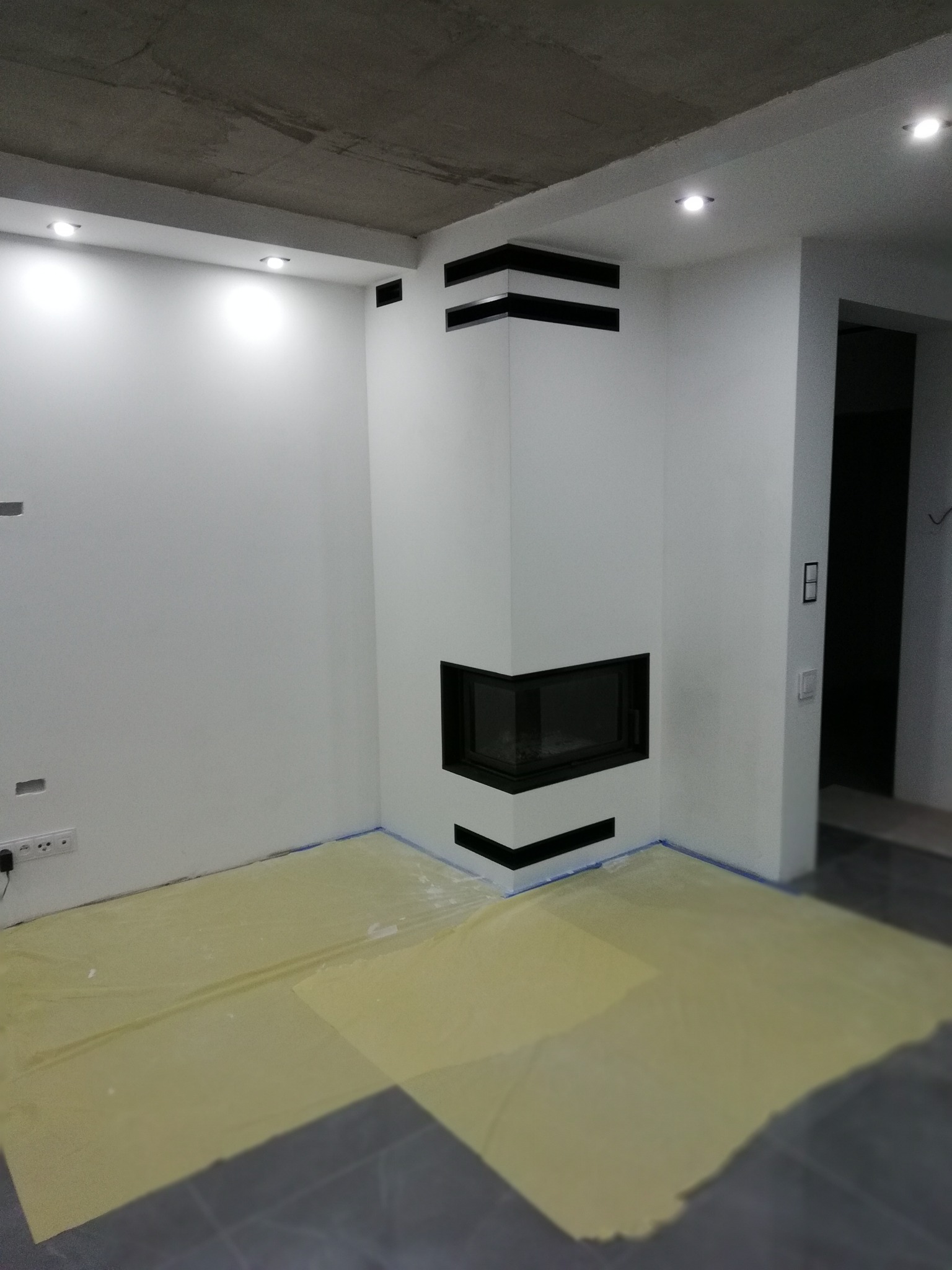 Wnętrze w trakcie wykańczania z narożnym, białym wkładem kominkowym z czarnymi akcentami, zabezpieczona folią podłoga i surowy betonowy sufit z halogenami.