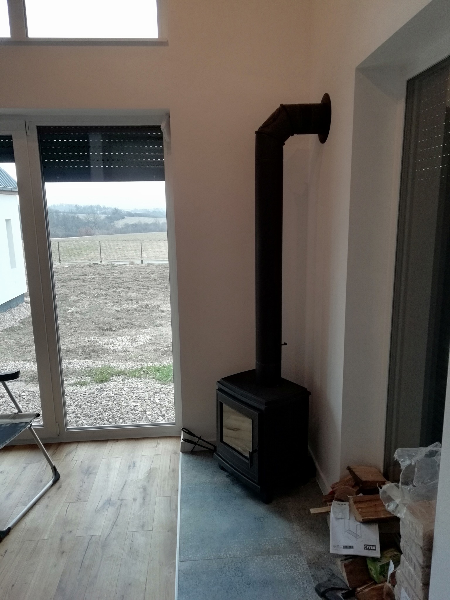 Czarny piec wolnostojący Koza K11 z rurą odprowadzającą dym, ustawiony w rogu pokoju z widokiem na zewnątrz przez duże okno. Obok pieca ułożone drewno opałowe i instrukcja obsługi.