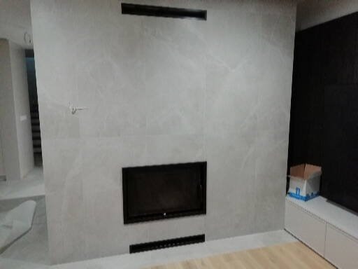 Zamontowany, nowoczesny kominek elektryczny obudowany płytami imitującymi marmur, z czarną ramą, widoczna górna i dolna kratka wentylacyjna, w tle fragment zabudowy meblowej i karton.