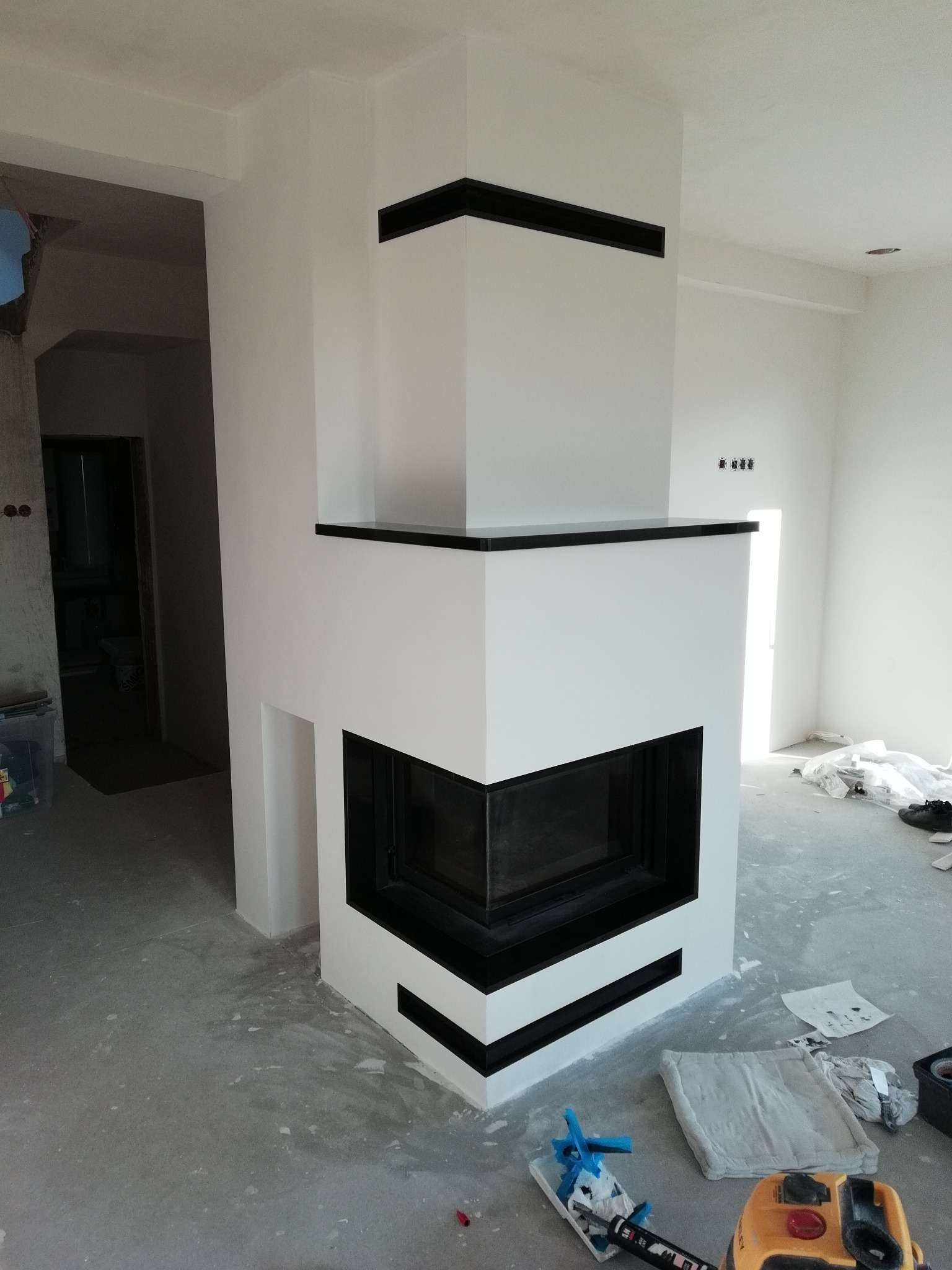 Nowoczesny, narożny wkład kominkowy z czarnymi akcentami, w trakcie montażu w surowym wnętrzu. Widoczne narzędzia malarskie i pozostałości po remoncie na podłodze.