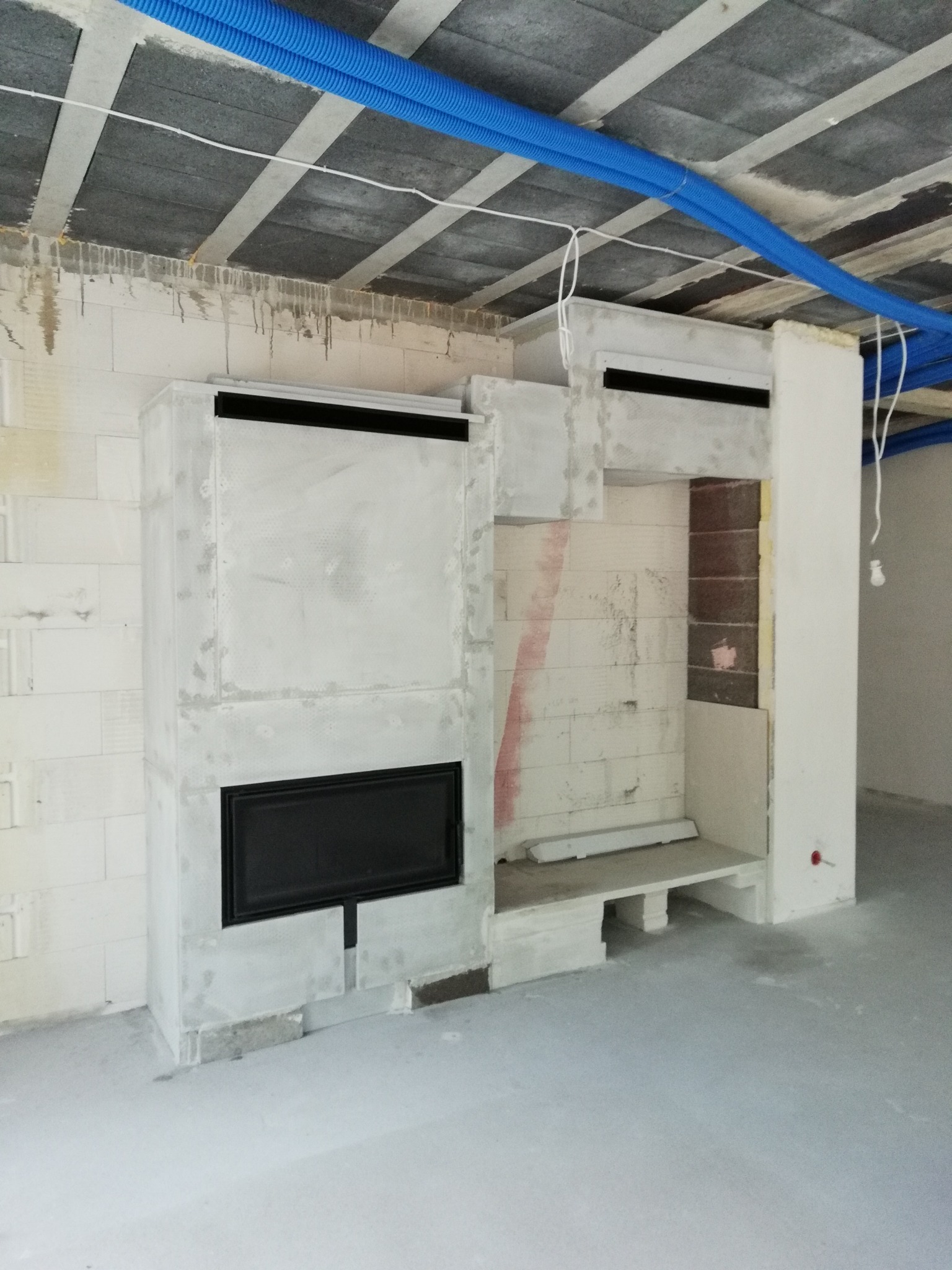 Surowy kominek z czarnym wkładem, w trakcie budowy, widoczne bloczki betonowe, niebieskie rury instalacyjne pod sufitem.