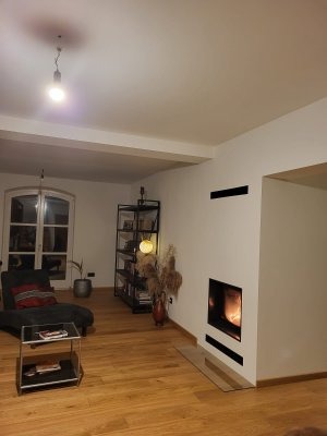 Nowoczesny salon z minimalistycznym wkładem kominkowym w zabudowie, drewnianą podłogą i czarnym regałem na książki.