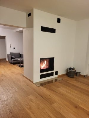 Nowoczesny kominek z paleniskiem otoczonym czarną ramą, wbudowany w białą ścianę z minimalistycznymi wlotami powietrza, uzupełniony o drewnianą podłogę i widok na szarą sofę w tle.