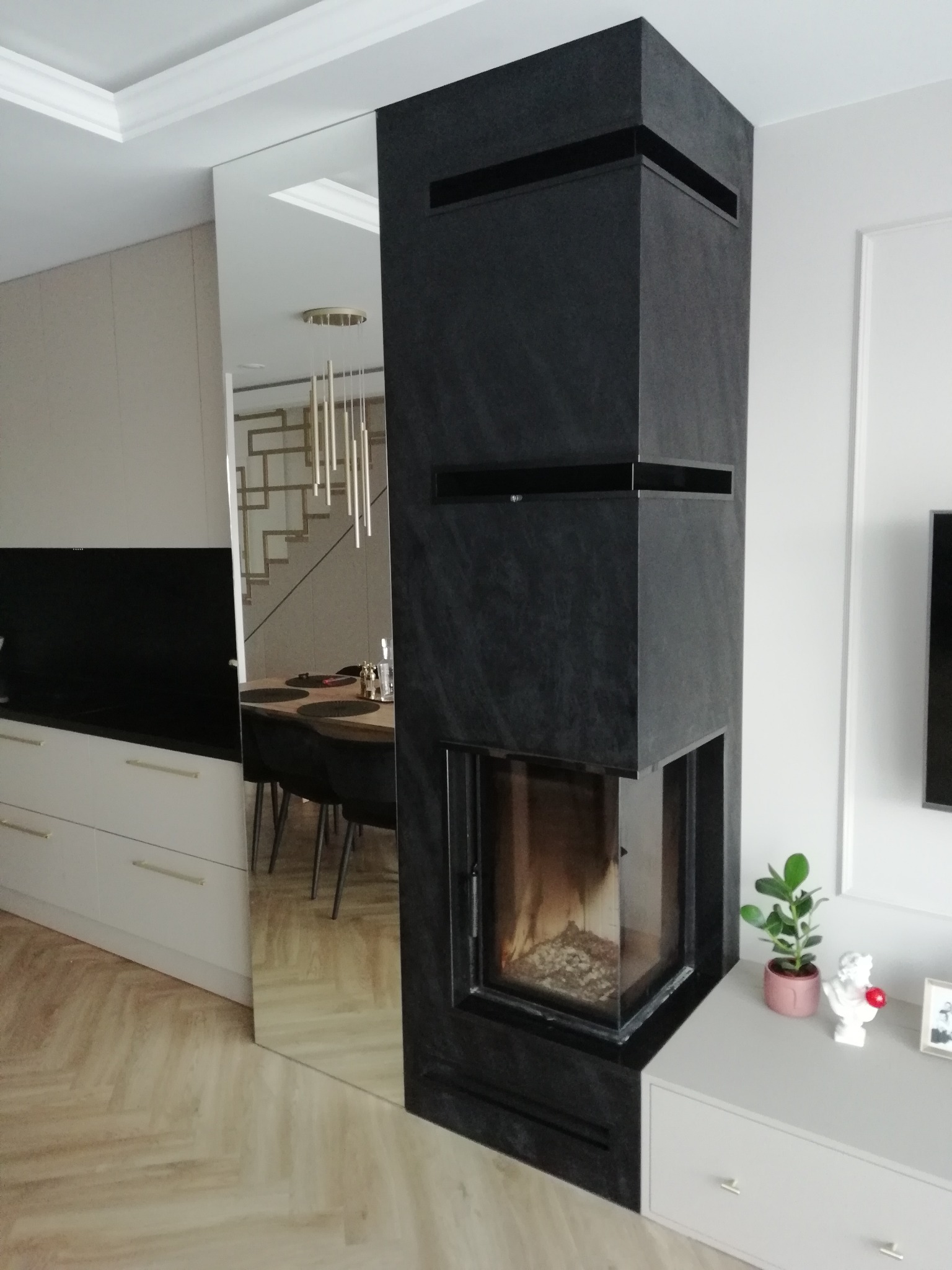 Nowoczesny, narożny piec obudowany czarnym kamieniem, z widocznym paleniskiem, wkomponowany w jasne wnętrze z lustrzaną ścianą i elementami dekoracyjnymi.