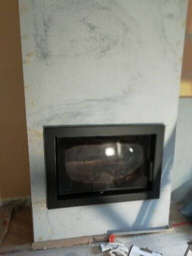 Wnętrze kominka z czarną ramą, obudowane płytami imitującymi marmur, widoczne ślady prac wykończeniowych na podłodze.