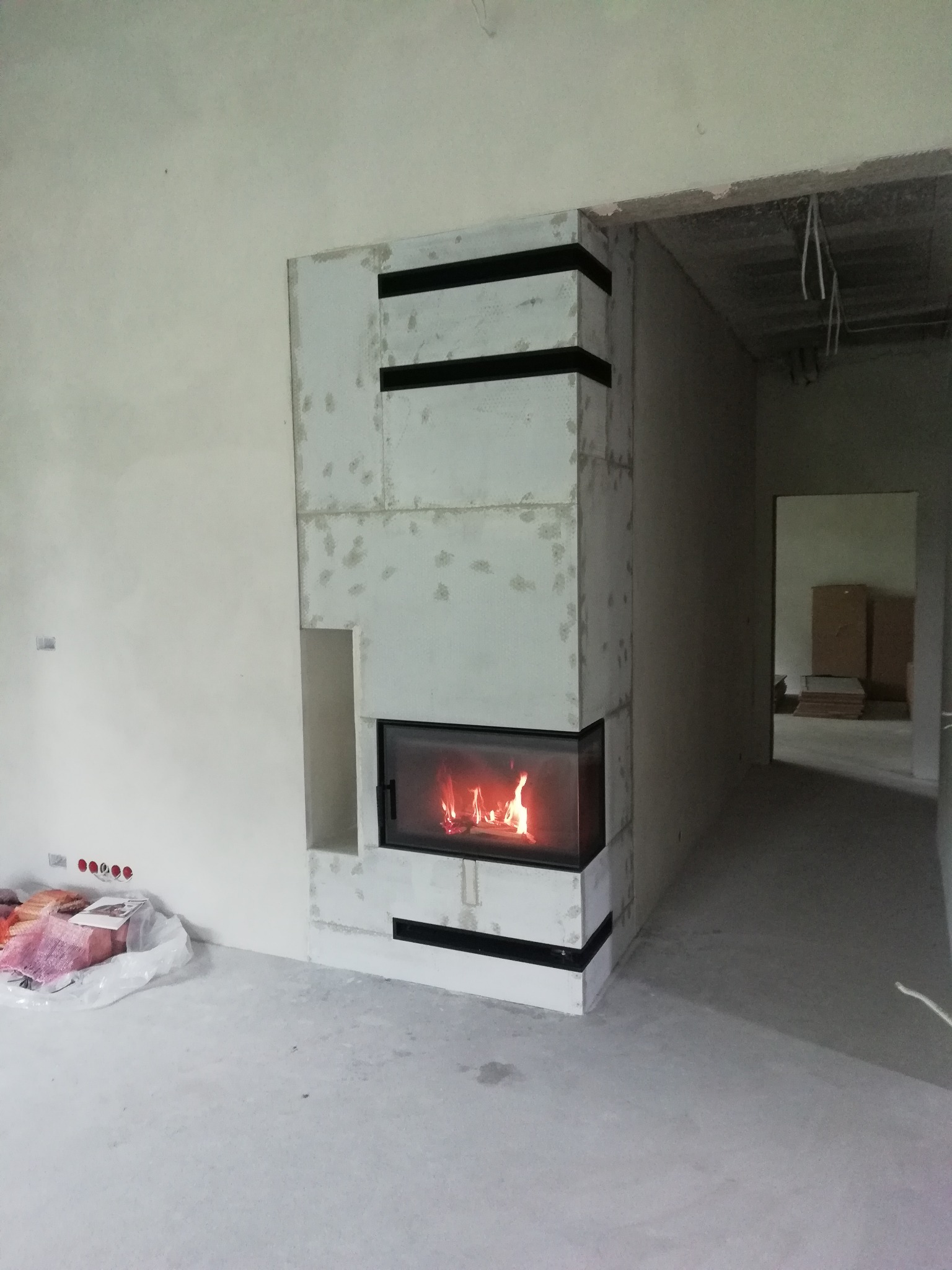 Nowoczesny kominek z paleniskiem w surowym wnętrzu. Widoczne elementy wykończeniowe i instalacja elektryczna w ścianie. Minimalistyczny design.