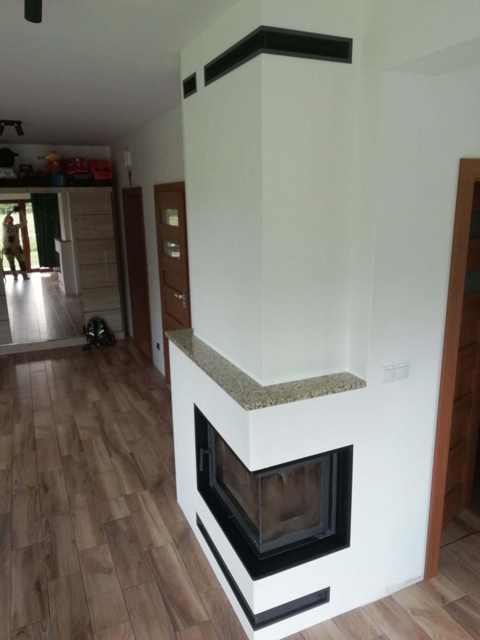 Nowoczesny kominek narożny z czarną obudową i blatem z jasnego kamienia, w jasnym salonie z drewnianą podłogą, widok z perspektywy podłogi.