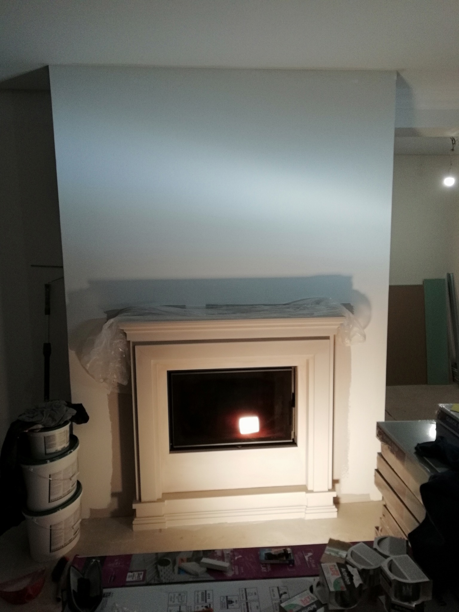 Biały kominek z paleniskiem w trakcie montażu, zabezpieczony folią, widoczne farby i narzędzia, ściana w jasnym odcieniu błękitu.