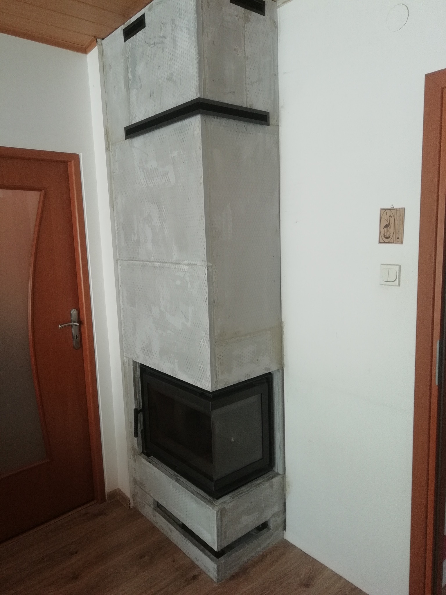 Narożny kominek z szarą obudową, czarnym wkładem i elementami dekoracyjnymi, umieszczony w rogu pomieszczenia z drewnianą podłogą i drzwiami.