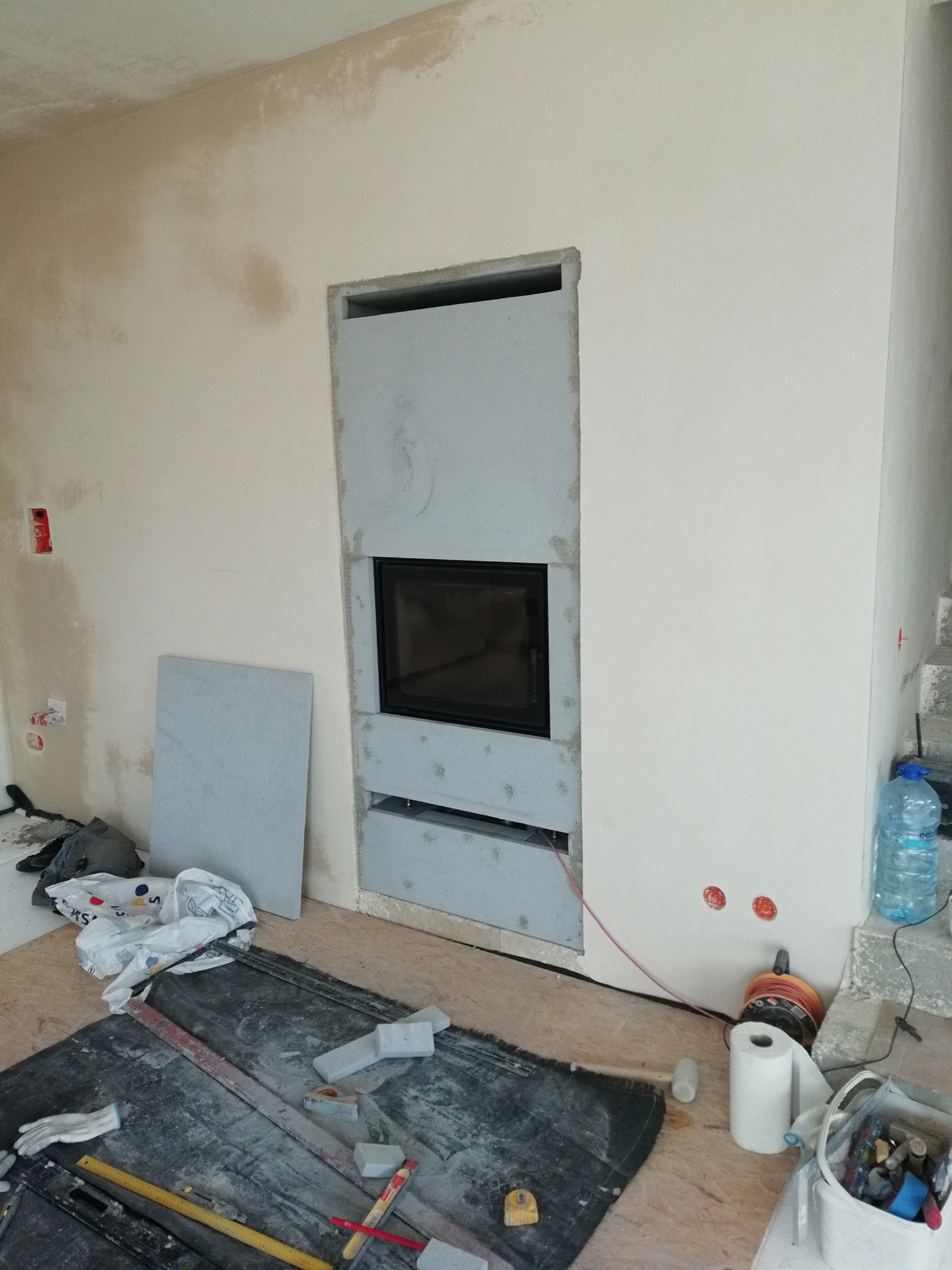 Wnętrze w trakcie montażu wkładu kominkowego, widoczne narzędzia i materiały budowlane, ściany w stanie surowym, wstępny etap obudowy kominka.