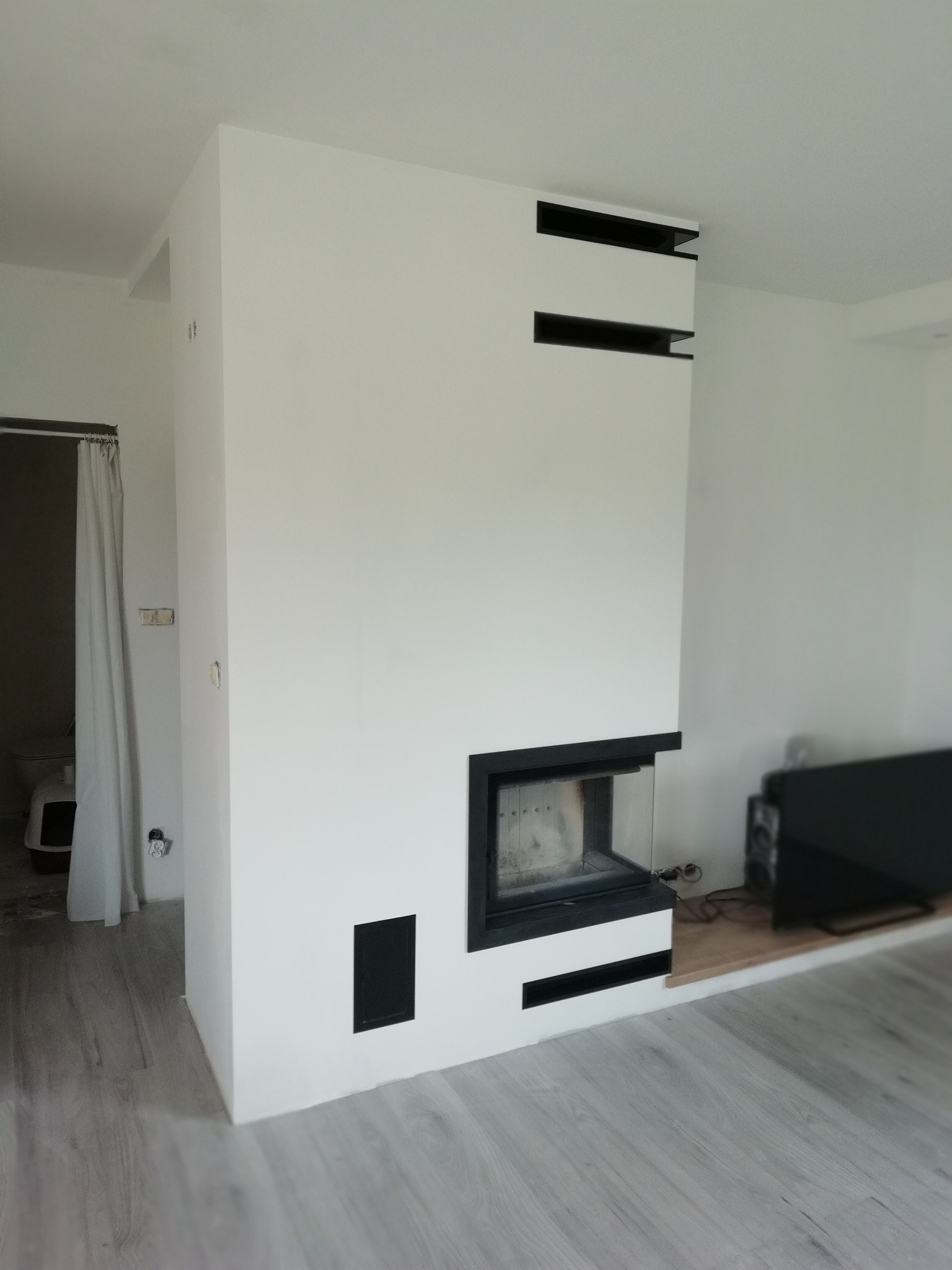 Nowoczesny kominek z czarnymi akcentami w salonie w Jedlinie-Zdroju. Minimalistyczny design, jasne ściany, widoczny fragment podłogi i telewizor w tle.