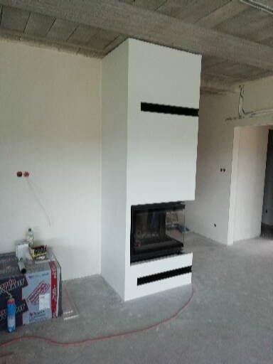 Nowoczesny, biały kominek z czarnymi detalami w surowym wnętrzu. Widoczne materiały budowlane sugerują etap wykończenia. Minimalistyczny design.