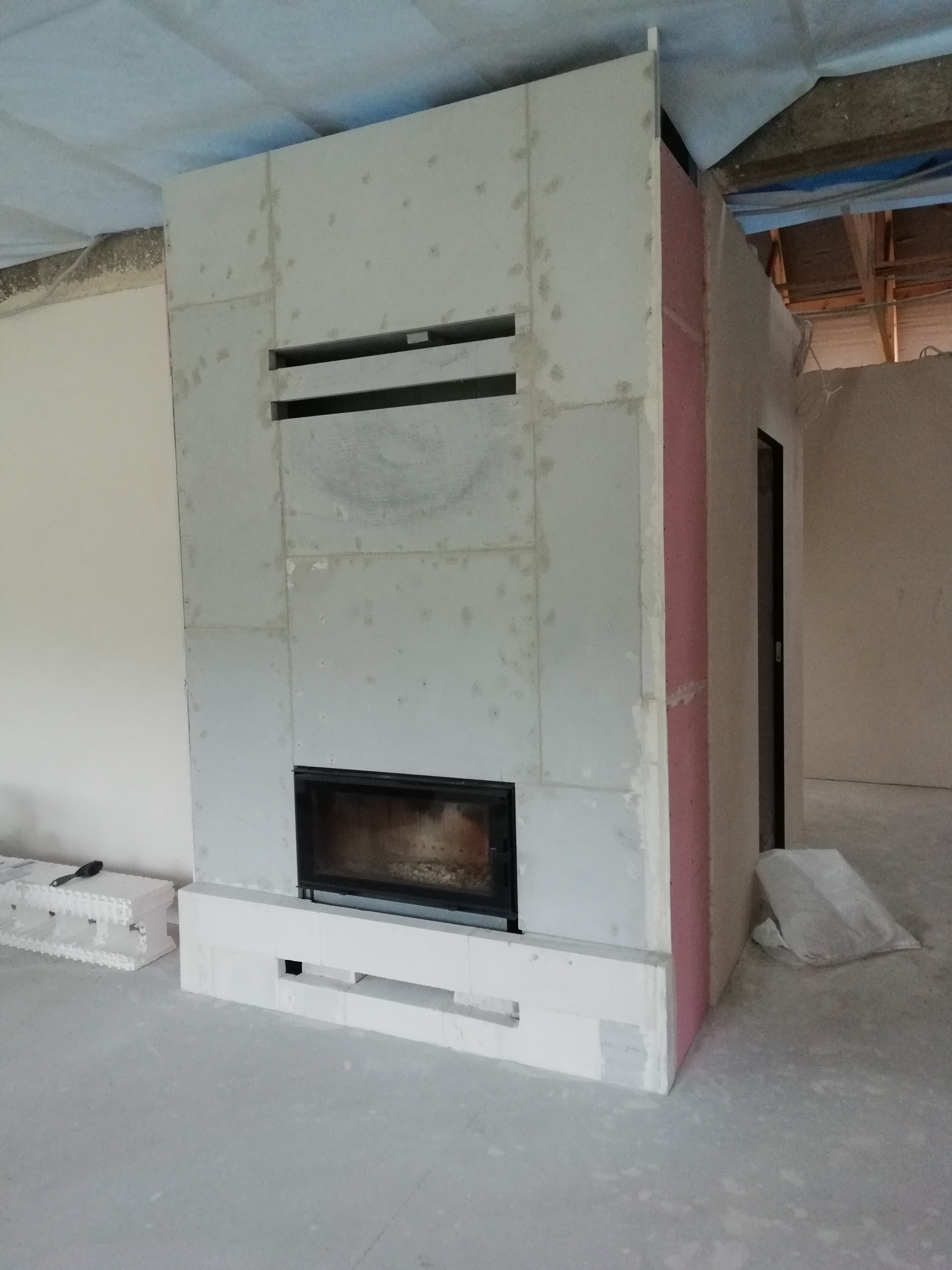Wnętrze w trakcie budowy z widocznym, częściowo obudowanym wkładem kominkowym z czarną ramą, obudowanym płytami gipsowymi i elementami izolacji. Widoczne materiały budowlane.