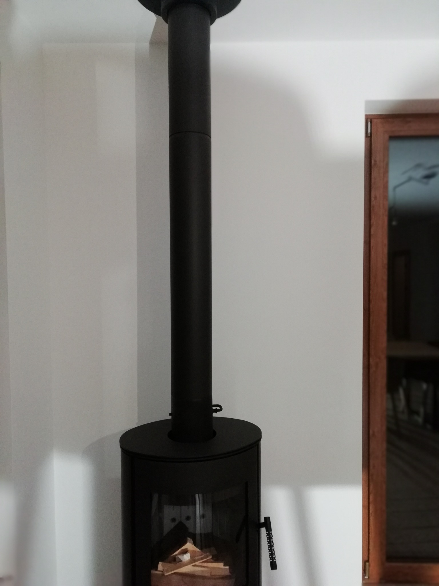 Czarny, owalny piec wolnostojący z rurą odprowadzającą dym, widoczny na tle białej ściany i drewnianej ramy drzwi. Wewnątrz paleniska ułożone drewno.