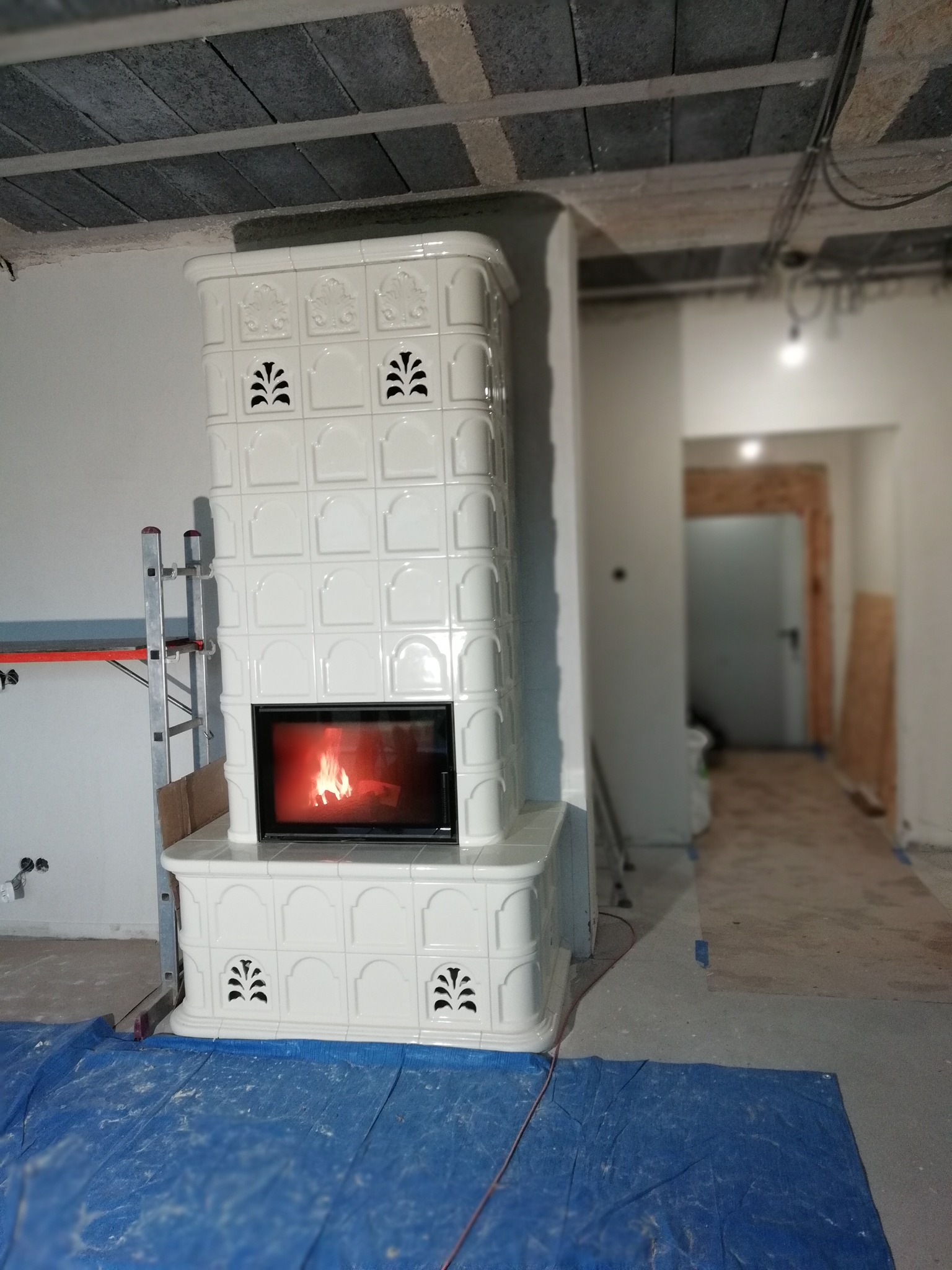 Biały, kaflowy piec kominkowy z paleniskiem, w surowym wnętrzu. Widoczna drabina i zabezpieczona podłoga. Płomienie w palenisku.
