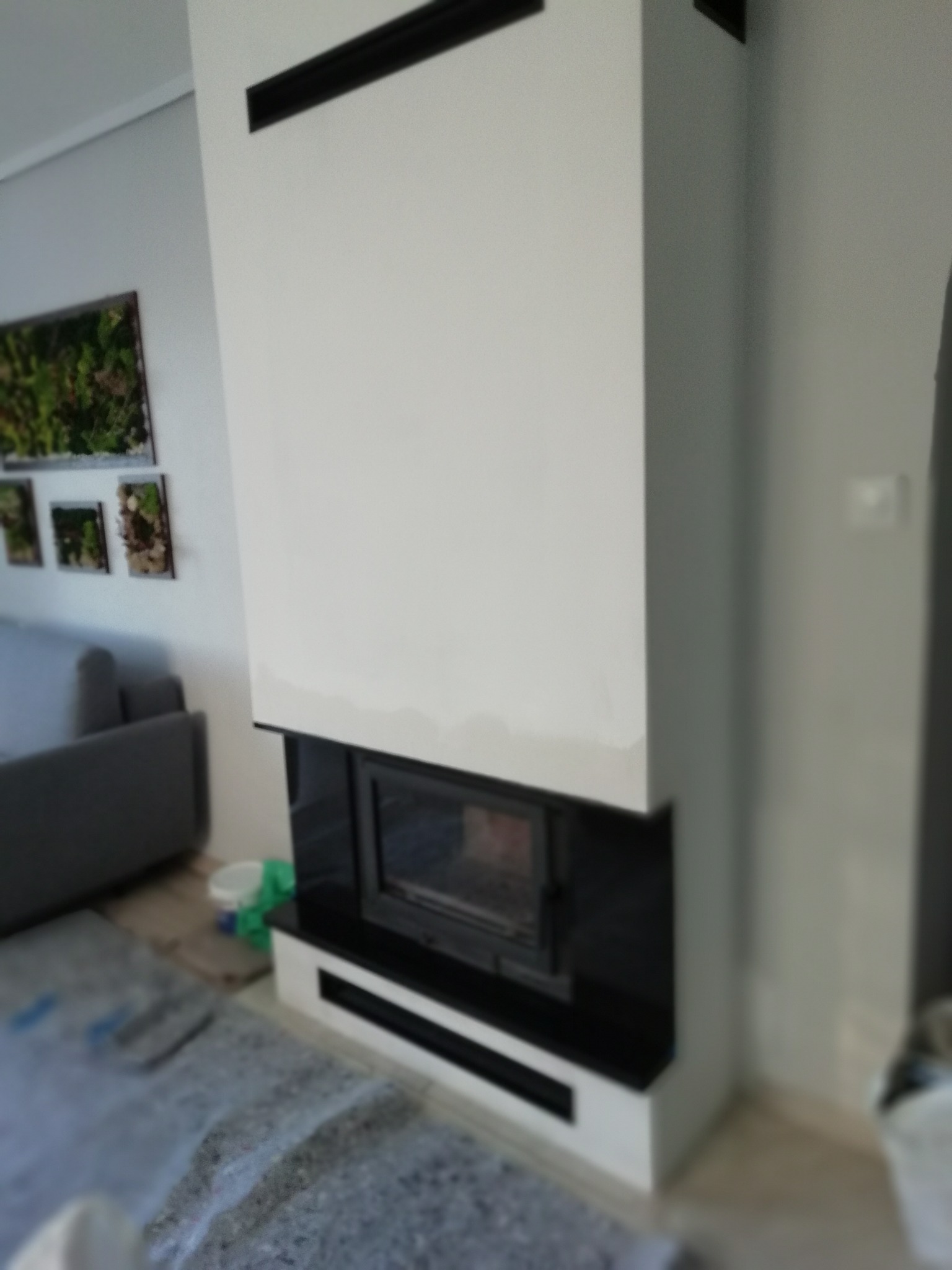 Biały kominek z czarnymi elementami, nowoczesny design. Widoczne dekoracje ścienne i fragment sofy. Na podłodze zabezpieczenie podczas prac.